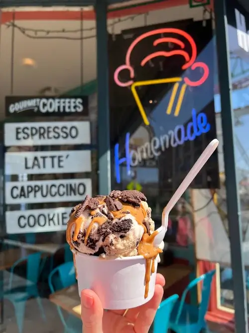 Hand holding a cup of cookies-and-cream ice cream topped with cookie pieces and caramel drizzle in front of an ice cream shop offering espresso, lattes, and cappuccinos.