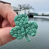 Small crocheted green four-leaf clover held between fingers with a waterfront and docked boats blurred in the background.