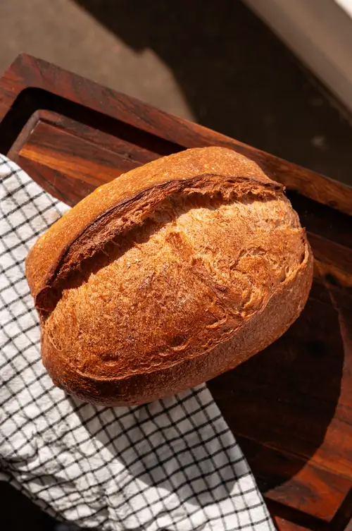 Freshly baked artisan sourdough bread loaf with a crisp golden-brown crust resting on a wooden cutting board beside a kitchen towel.